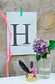 Auf Papier gedruckter Buchstabe H an Betonwand, davor Hortensie in Cocktail-Shaker, Feder und pink gefärbter Ast