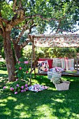 Different patterned scatter cushions on rustic garden swing seat under shady tree with picnic basket on ground