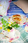 Mug of yellow wildflowers in front of floral plate of focaccia sandwiches on picnic blanket on bed