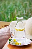 Carafe filled with water and slices of lemon and wild flowers on plate in front of straw hat on wooden surface