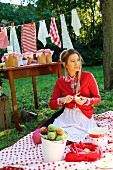 Junge Frau auf Picknickdecke sitzend schält Äpfel, im Hintergrund Tisch mit Einmachgläsern und Wäscheleine