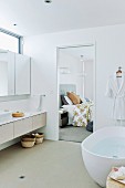 Free-standing bathtub and mirrored cabinet above continuous washstand with drawers next to door of ensuite bathroom with view into bedroom