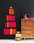 Stylised Christmas tree made from neon woollen fringes on black wall, arrangement of vases on retro sideboard and stacked presents on floor