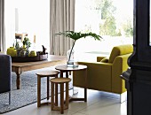 Set of wooden side tables next to yellow armchair, tray and various yellow vases on rustic wooden coffee table and view into garden through glass wall