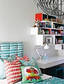 Verschieden gemusterte Kissen auf Bett neben weißem Schreibtisch mit Tischleuchte, an Wand montierte Regalmodule in Weiß und Rot
