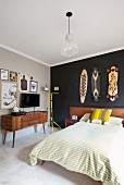 Französisches Bett mit Holz Kopfteil vor schwarz getönter Wand mit aufgehängten Skateboards, an der Seite Fifty Sideboard vor hellgrau getönter Wand mit Fernseher zwischen Zeichnungen
