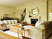 Pale sofa set with arranged scatter cushions and leather-covered ottoman in front of open fireplace in grand living room