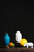 Arrangement of white head of Buddha, white hippo ornament, fruits and blue-glazed urn against dark background