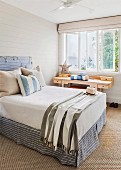 Scatter cushions on double bed with headboard against pale grey wooden wall next to wooden desk below window in simple bedroom