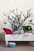Romantic arrangement of pale blue, bird-shaped decorations hanging on spring branches and pink bellis in zinc pot in white planter