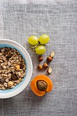 A bowl of muesli next to grapes, nuts and an apricot