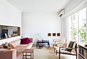Minimalistischer, weisser Wohnraum - Pastellrosa Polstercouch und Beistelltisch gegenüber Sessel vor Fensterfront, Wandausschnitt mit Blick ins Arbeitszimmer