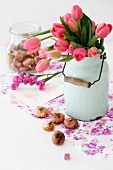 Zart roter Tulpenstrauß in nostalgischer weißer Milchkanne und Blumenzwiebeln auf lilafarbenem Geschenkpapier
