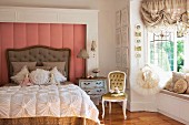 Romantic scatter cushions and ornate bedspread on antique bed against wall panel with dusky pink cover next to tutu on clothes hanger in bay window with ruched curtains