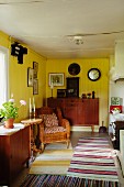 Rustic interior with yellow-painted walls, various rugs striped in different colours, rattan armchair and sideboard