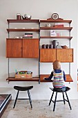 Little girl sitting on wooden stool in front of retro-style, modular shelving system with compartments, cabinets and writing desk on wall-mounted rails