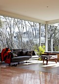 Modern sofa and rustic side table in interior with glass walls and view of trees