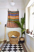 Ethnic wall hanging above wicker armchair and potted palm tree on diagonal chequered floor