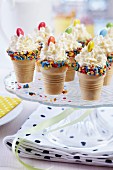Cones of ice cream edged with sugar confetti and decorated with cream and smarties festively arranged on cake stand