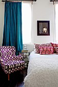 Armchair and scatter cushions with covers in various retro patterns and pale bedspread on bed