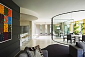 Lounge area with black wall in front of light-flooded dining area behind curved glass partition in spacious modern interior