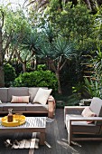 Grey-upholstered outdoor furniture and coffee table on wooden terrace with yuccas in mature garden in background