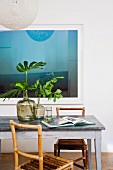 Blue reflective artwork behind vintage table with metal top, bamboo chairs and green leaves arranged in demijohns