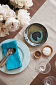 Cutlery on blue napkin on plate next to bowl with peacock-heather motif and small wooden bowls on pale linen runner