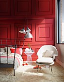 Metall-Bettgestelll in Anthrazit, weisser Sessel mit passendem Fussschemel auf weißem Flokati Teppich vor rot lackierter Holz- Kassettenwand