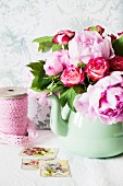 A bunch of peonies and roses in an old-fashioned green metal jug on a shelf in front of a wallpapered wall
