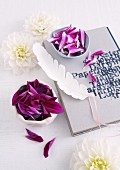 White dahlia flowers, china bowls of purple petals and white feather on diary with printed cover