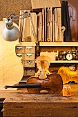 Unbespannte Geige auf rustikaler Hobelbank mit anderen Geigen-Modellen und sortierten Hölzern auf Regalbrett, beleuchtet mit Retro-Schreibtischlampe