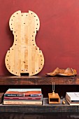 Rustikale Holzablage mit Bücherstapel und Stimmgabel, Schuh-Modelle aus Holz und heller Cellokorpus an dunkelrote Wand gelehnt