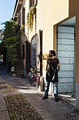 Italienischer Geigenbauer mit Arbeitsschürze an Hauswand vor Werkstatt gelehnt