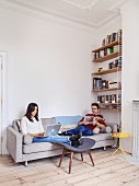 Young couple relaxing with magazine and laptop on grey sofa in renovated period apartment with wooden floor, bookcase and stucco ceiling