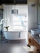Modernes Bad - Seitlich in Nische eingebauter Waschtisch mit Unterschrank aus Holz, freistehende Badewanne vor geschlossenem, transparentem Vorhang am Fenster