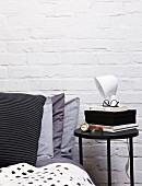 Black side table and stacked scatter cushions in shades of grey on bed against whitewashed brick wall
