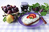 Bread with cottage cheese on a purple plate next to a basket of plums, an apple and plum jam