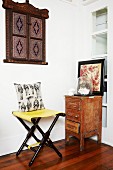 Folding stool in corner next to small, vintage cabinet & wooden objet d'art on wall