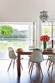 Classic, white shell chairs around wooden table in modern interior in front of open terrace door with view of lake