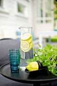 Glass carafe of lemon water, blue drinking glasses and pot of herbs on black side table