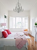 Double bed with pink scatter cushions on white bed linen below Rococo chandelier in white, wood-clad bedroom