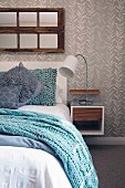Light blue knitted blanket on bed with scatter cushions in various shades of blue and bedside cabinet with table lamp mounted on wall with grey and white wallpaper