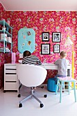 Girl and boy seated at desk in retro child's bedroom with pink floral wallpaper