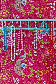 Hakenleiste mit Halsketten aus farbigen Perlen an Wand aufgehängt, Tapete mit Blumenmuster auf pinkfarbenem Hintergrund