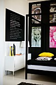 Detail of four-poster bed with black metal frame and yellow and patterned scatter cushions below collection of posters on black wall
