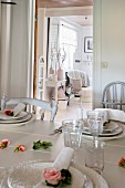 Place settings and roses on white dining table; view into rustic living room through open door in background