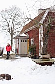 Woman and dog in snowy garden in front of falu red wooden house with porch