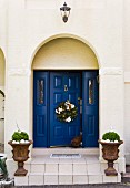 Christmas wreath hung on blue wooden panelled door with planted urns on front steps