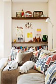 Two white dogs and many scatter cushions with graphic patterns on corner sofa in front of bookshelves in niche